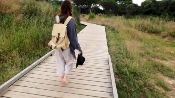 Wandern. Ein Mädchen mit Rucksack geht auf einem Küstenweg. Ein Mädchen mit langen Haaren läuft an einer Uferpromenade entlang. Sonniger Sommertag. Malerische Straße. Urlaub am Meer. Der Pfad im Schilf. — Stockvideo