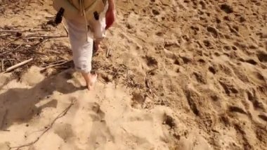 Yürüyüş. Sırt çantalı çıplak ayaklı kız kumsalda yürüyor. Uzun saçlı ve siyah şapkalı bir kız kıyı boyunca yürüyor. Güneşli bir yaz günü. Resimli deniz körfezi boyunca yürü. Denizde tatil..