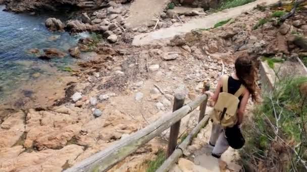 Caminhadas. Uma menina com uma mochila caminha em um caminho costeiro. Uma menina com cabelos longos está andando ao longo de uma costa rochosa. Dia de verão ensolarado. Caminhe ao longo da pitoresca baía do mar. Férias no mar. Baía desabitada — Vídeo de Stock