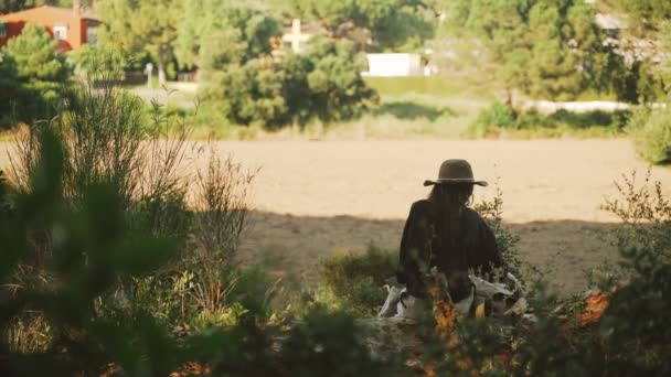 Una joven está leyendo un libro en el bosque. Una chica con una camisa negra y un sombrero de paja se sienta en un árbol caído y hojea un libro. Vacaciones en el pueblo. Día soleado de verano. El viento sopla pelo largo . — Vídeos de Stock