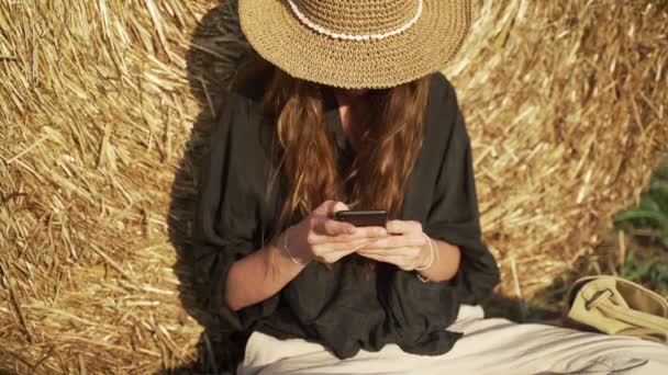 Una giovane donna che messaggia su uno smartphone sul campo. Una ragazza con una camicia nera e un cappello di paglia seduta appoggiata a un pagliaio a guardare il telefono. Vacanza nel villaggio. Giornata estiva soleggiata. Vacanze. — Video Stock