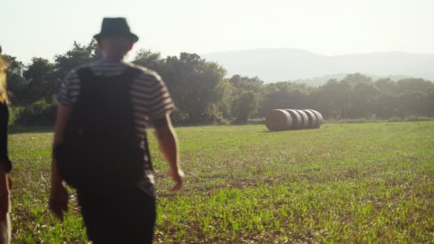 Young couple walks along a beautiful field among the forest and mountains at sunset. Man and woman in a straw hats walks on a sloping field with stacks. Vacation in the village. Golden evening. Love. — Stock Video