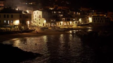 Geceleri güzel bir sahil köyü manzarası. Küçük Akdeniz kasabası. Kumsaldaki beyaz evler. Sahilde tatiller. Pencerelerdeki ışık denizin dalgalarına yansıyor..