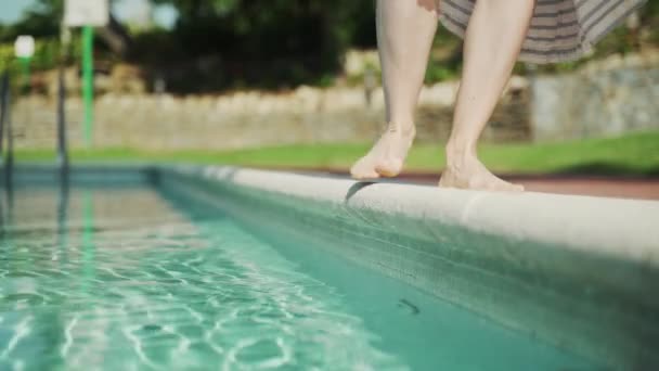 Ragazza a piedi nudi in un abito a righe sta camminando lungo il bordo della piscina e assaggia l'acqua con il piede. Acqua dolce turchese in piscina in una giornata calda. Vacanze estive. Giornata estiva soleggiata. — Video Stock