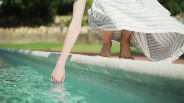 Barefoot girl in a striped dress is sitting on the edge of the pool and tastes the water with her hand. Turquoise fresh water in the pool on a hot day. Summer holidays. Sunny summer day. — Stock Video