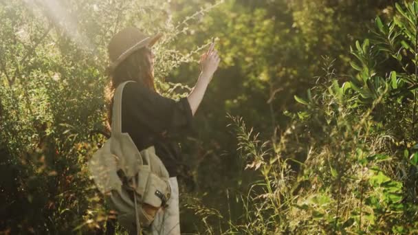 Eine junge Frau fotografiert mit ihrem Smartphone Bäume im Wald. Ein Mädchen in schwarzem Hemd und Strohhut filmt den Wald. Urlaub im Dorf. Sonniger Sommertag. Wind weht lange Haare. — Stockvideo