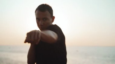 Sabah sporu. Deniz rıhtımında Muay Thai Kutusu eğitimi. Delikanlı denizin arka planına ve doğan güneşe karşı yumruk pratiği yapıyor. Spor aktivitesi. Sporcu. Yumruk, dirsek. Yumruk pratiği.