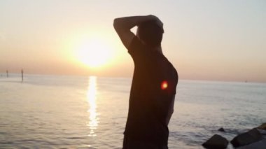 Sabah sporu. Deniz rıhtımında antrenmandan önce ısınma ve esneme hareketleri. Genç adam denizin arka planına ve doğan güneşe karşı pratik yapıyor. Spor aktivitesi. Atlet silueti. Mangaları atlatır..