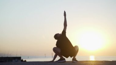 Sabah sporu. Deniz rıhtımında antrenmandan önce ısınma ve esneme hareketleri. Denizin ve güneşin doğuşuna karşı seni çalıştıran genç adam. Spor aktivitesi. Atlet silueti. 
