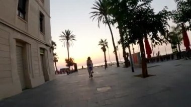 Elbiseli ve sırt çantalı bir kız güzel bir set boyunca bisiklet sürüyor. Deniz boyunca bisiklet sürüyor. Sabah Şehri uyanıyor. Doğan güneşin arka planında ağaçlar ve palmiyeler var..