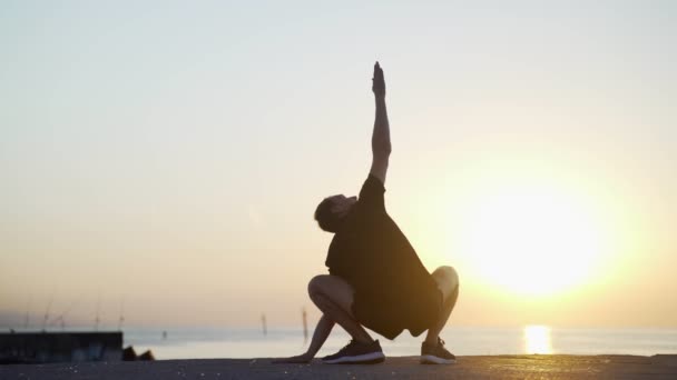 Sabah sporu. Deniz rıhtımında antrenmandan önce ısınma ve esneme hareketleri. Denizin ve güneşin doğuşuna karşı seni çalıştıran genç adam. Spor aktivitesi. Atlet silueti. — Stok video