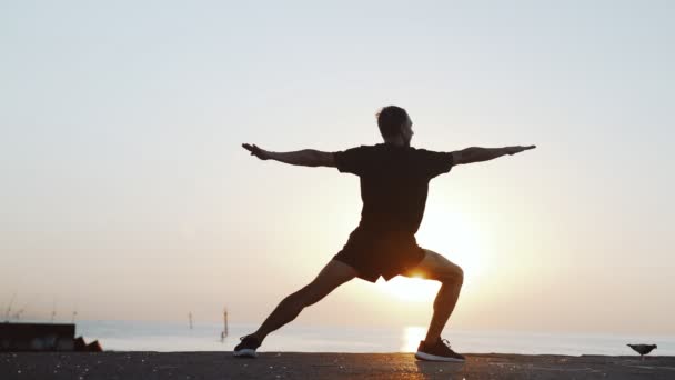Entraînement matinal. Réchauffez-vous et étirez-vous avant de vous entraîner sur la jetée. Jeune homme pratiquant youga dans le contexte de la mer et le soleil levant. Activité sportive. Silhouette d'athlète. — Video