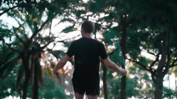 Entrenamiento matutino. Un joven saltando la cuerda en el soleado parque urbano. Actividad deportiva. Atleta. Ejercicios en la ciudad. Hombre en camiseta negra entrenando entre árboles y palmeras. Estilo de vida saludable. — Vídeo de stock