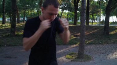 Sabah sporu. Parkta Muay Thai Kutusu eğitimi. Genç adam güneşli şehir parkında koşuyor ve yumruk pratiği yapıyor. Spor aktivitesi. Sporcu. Yumruk, dirsek, diz. Yumruk antrenmanına yakın çekim.