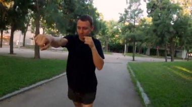 Sabah sporu. Parkta Muay Thai Kutusu eğitimi. Genç adam güneşli şehir parkında koşuyor ve yumruk ve tekme pratiği yapıyor. Spor aktivitesi. Sporcu. Zıplıyor ve koşuyor. Yumruk, dirsek, diz.