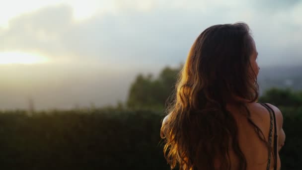 Piękny letni wieczór. Dziewczyna w zielonej sukience siedzi na tarasie i patrzy na piękny zachód słońca w górach. Letni spokój. Złote światło pośród chmur. Romantyczny wieczór. Młoda kobieta. — Wideo stockowe