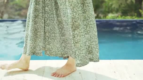 Barefoot meisje in een groene jurk loopt langs de rand van het zwembad. Turkoois zoet water in het zwembad op een warme dag. Lichte rimpelingen op het wateroppervlak. Zomervakantie. Zonnige zomerdag. — Stockvideo