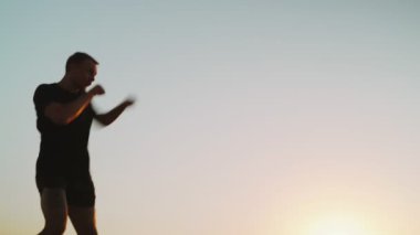 Sabah sporu. Deniz rıhtımında Muay Thai Kutusu eğitimi. Denizin arka planına ve yükselen güneşe karşı tekme pratiği yapan genç adam. Spor aktivitesi. Sporcu. Yumruk, dirsek. Yumruk antrenmanına yakın çekim