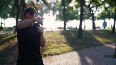 Sabah sporu. Parkta Muay Thai Kutusu eğitimi. Genç adam güneşli şehir parkında koşuyor ve yumruk pratiği yapıyor. Spor aktivitesi. Sporcu. Yumruk, dirsek, diz. Yumruk antrenmanına yakın çekim.