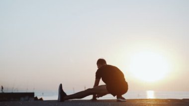 Sabah sporu. Deniz rıhtımında antrenmandan önce ısınma ve esneme hareketleri. Genç adam denizin arka planına ve doğan güneşe karşı pratik yapıyor. Spor aktivitesi. Atlet silueti. Mangaları atlatır..