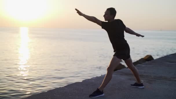 Entrenamiento matutino. Caliente y estire antes de entrenar en el muelle del mar. Joven practicando youga contra el telón de fondo del mar y el sol naciente. Actividad deportiva. Silueta de atleta. — Vídeos de Stock