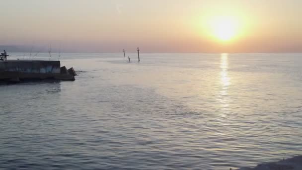 Slunce vychází na mořském molu. Klidné ranní moře. Rybářské pruty na molu. Jasné slunce na barevné obloze. Klidné ráno u moře. — Stock video