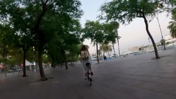 Girl in a dress and with a backpack rides a bicycle along a beautiful embankment. Cycling along the sea. Morning city waking up. Coast with trees and palms against the background of the rising sun. — Stock Video