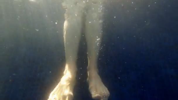 Meisje in een zwarte bikini zit aan de rand van het zwembad en bungelt haar benen. Schieten onder water. Verfrissend zwembad in warme zomer. Spatten en bellen in blauw water. Zomer chill. Close-up van de voeten. — Stockvideo