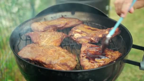 Barbacoa de verano con amigos. Costillas de cerdo asadas. Primer plano de un chefs manos cepillado costillas de cerdo con salsa barbacoa casera. Comida sabrosa. Comida al aire libre. La carne a la parrilla está fumando en la red de parrilla. — Vídeos de Stock