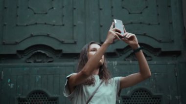 Güzel, uzun saçlı esmer kadın büyük bir kapının önünde fotoğraf çekiyor. Çekici bir kadın çarpıcı binaların fotoğraflarını çekiyor. Güzel bir şehirde güzel bir turist selfie çekiyor. Cep telefonu..