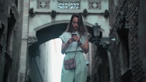 Mooie langharige brunette controleert haar mobieltje onder de boog. Jonge aantrekkelijke vrouw die sms 't in prachtige gebouwen. Mooie toeristische wandelingen in een prachtige stad. Meisje met mobiele telefoon — Stockvideo