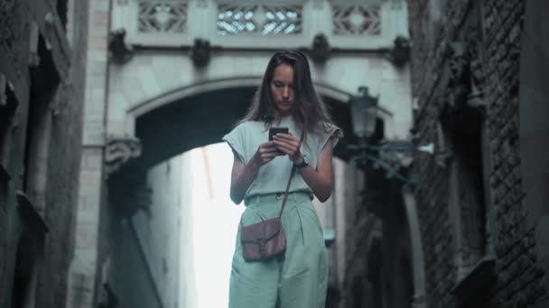 Hermosa morena de pelo largo comprueba su teléfono celular debajo del arco. Mujer atractiva joven mensaje de texto en edificios impresionantes. Bonitos paseos turísticos en una hermosa ciudad. Chica con teléfono móvil — Vídeos de Stock