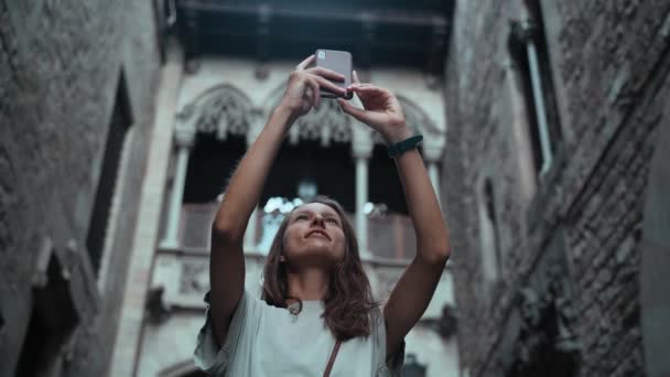 Linda morena de cabelos longos fazendo foto sob o arco. Jovem mulher atraente tirando fotos de edifícios deslumbrantes. Bonito turista faz selfie em uma bela cidade. Menina com telefone celular. — Vídeo de Stock