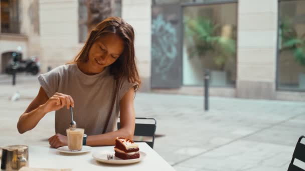 아름다운 머리의 여자가 카페에서 커피를 마시고 있습니다. 식당 테라스에 있는 젊은 여자가 카푸치노 거품을 먹고 있어요. 회색 티셔츠를 입은 갈색 머리 소녀가 작은 광장에 있는 카페에 앉아 있습니다. 아침 식사. — 비디오