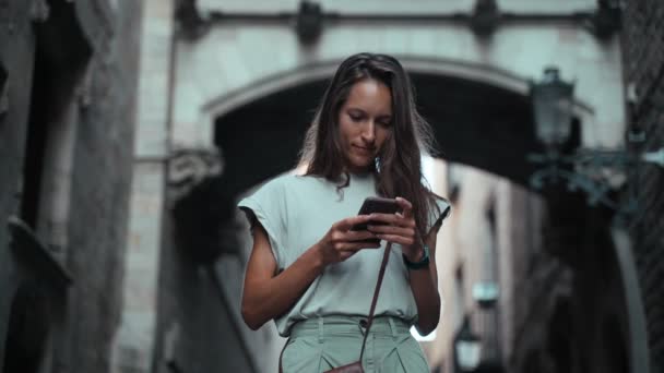 Beautiful long-haired brunette checks her cellphone under the arch. Young attractive woman texting message at stunning buildings. Pretty tourist walks in a beautiful city. Girl with mobile phone Royalty Free Stock Footage