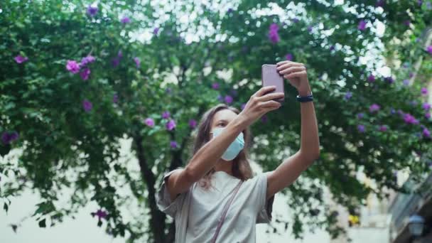 Mooie langharige brunette met beschermend masker maakt selfie op het plein met bloeiende bomen. Jonge aantrekkelijke vrouw die foto 's maakt in een prachtige stad. COVID-19 voorzorg. Blijf veilig. Coronavirus. — Stockvideo