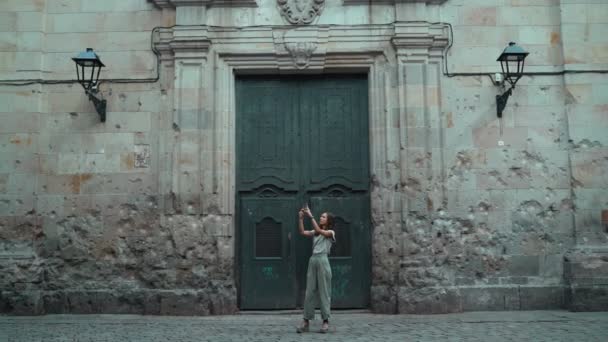 Piękna, długowłosa brunetka robiąca zdjęcie przed dużymi, rzeźbionymi drzwiami. Młoda atrakcyjna kobieta robi zdjęcia oszałamiającym budynkom. Pretty turysta robi selfie w pięknym mieście.Telefon komórkowy. — Wideo stockowe