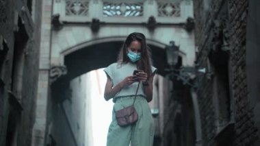 Koruyucu maskeli güzel, uzun saçlı esmer kemerin altındaki cep telefonunu kontrol ediyor. Sokakta mesaj atan çekici genç bir kadın. COVID-19 önlemi. Kendine dikkat et. Cep telefonlu turist