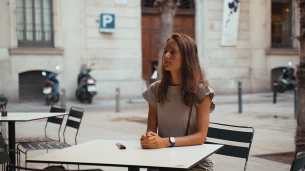 Hermosa mujer de pelo largo está esperando su orden en una mesa en un café. Mujer joven en la terraza de un restaurante. Chica de cabello castaño en una camiseta gris en un café en una pequeña plaza. Desayunando. — Vídeo de stock