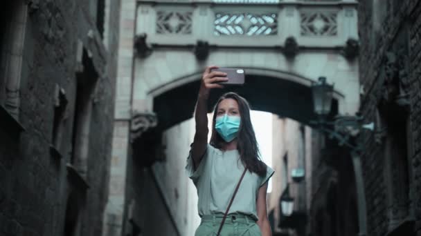 Bela morena de cabelos longos em máscara protetora faz selfie sob o arco. Jovem mulher atraente tirando fotos em uma bela cidade. Precaução COVID-19. Fica em segurança. Turista com telefone celular — Vídeo de Stock