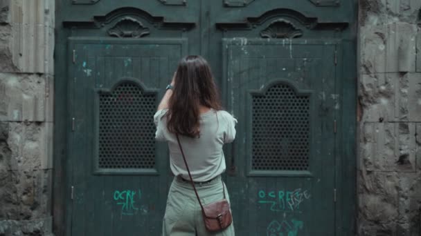 아름다운 긴 머리의 브라운 렛이 커다란 조각 된 문의 사진을 찍고 있습니다. 멋진 건물들의 사진을 찍고 있는 매력적 인 젊은 여성. 예쁜 관광객이 아름다운 도시에서 셀피를 만들고 있습니다. 모바일 폰. — 비디오