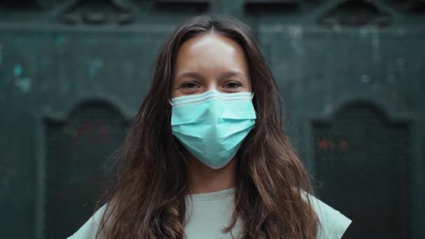 Mooie langharige brunette met beschermend masker. Jonge aantrekkelijke vrouw beschermt zichzelf tegen infectie. COVID-19 voorzorg. Blijf veilig. Coronavirus. Mooi meisje in een grijs t-shirt. — Stockvideo