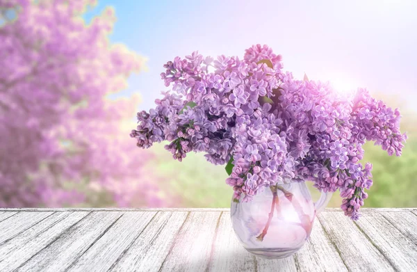 木桌在破旧别致的样式与丁香花在玻璃花瓶和春天树反对蓝色天空 — 图库照片