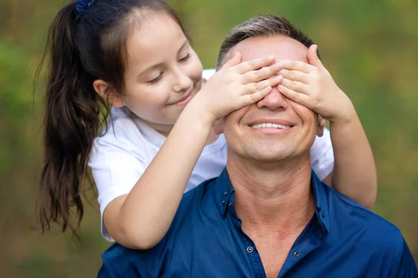 Cute little girl covering eyes of her father outdoors. Daughter makes surprise to her dad