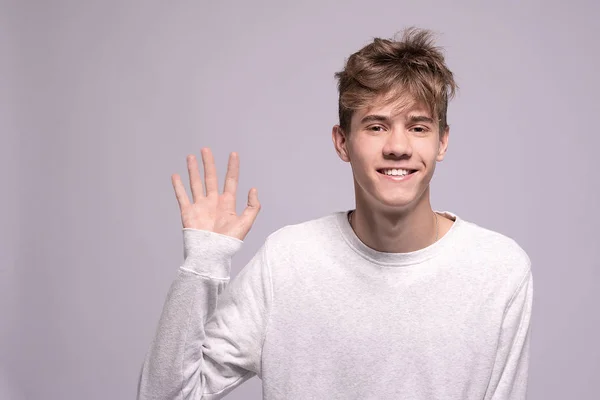 Young man smiling and saying hello at you over gray background. Portrait of handsome teenager guy 16-18 years old waving his hand