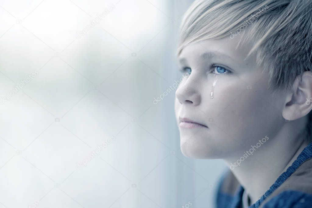 Crying boy looks out the window. Portrait of sad blond teen 12-14 year old 