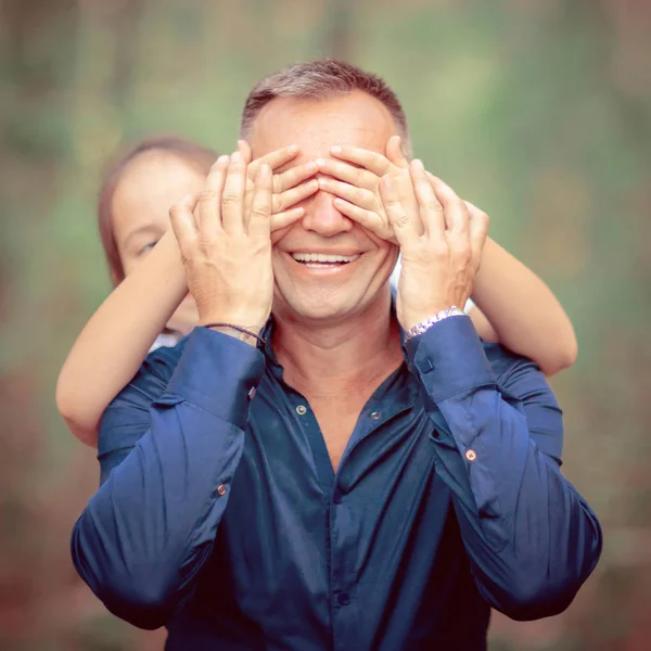 Cute little girl covering eyes of her father outdoors. Daughter makes surprise to her dad