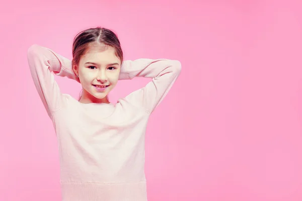 可爱的孩子女孩与手在头后面在一个轻松的姿势在粉红色的背景 — 图库照片