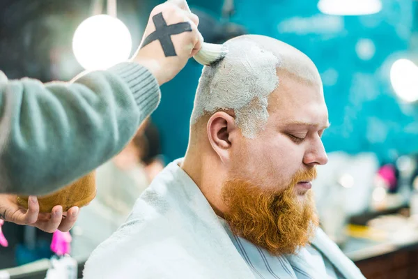 A redhead bearded man sitting in barbershop and have his head being creammed with shaving brush on.