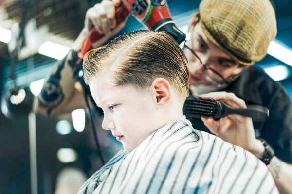 A hairdo for a boy is styling with a brush and hair dryer in barbershop.